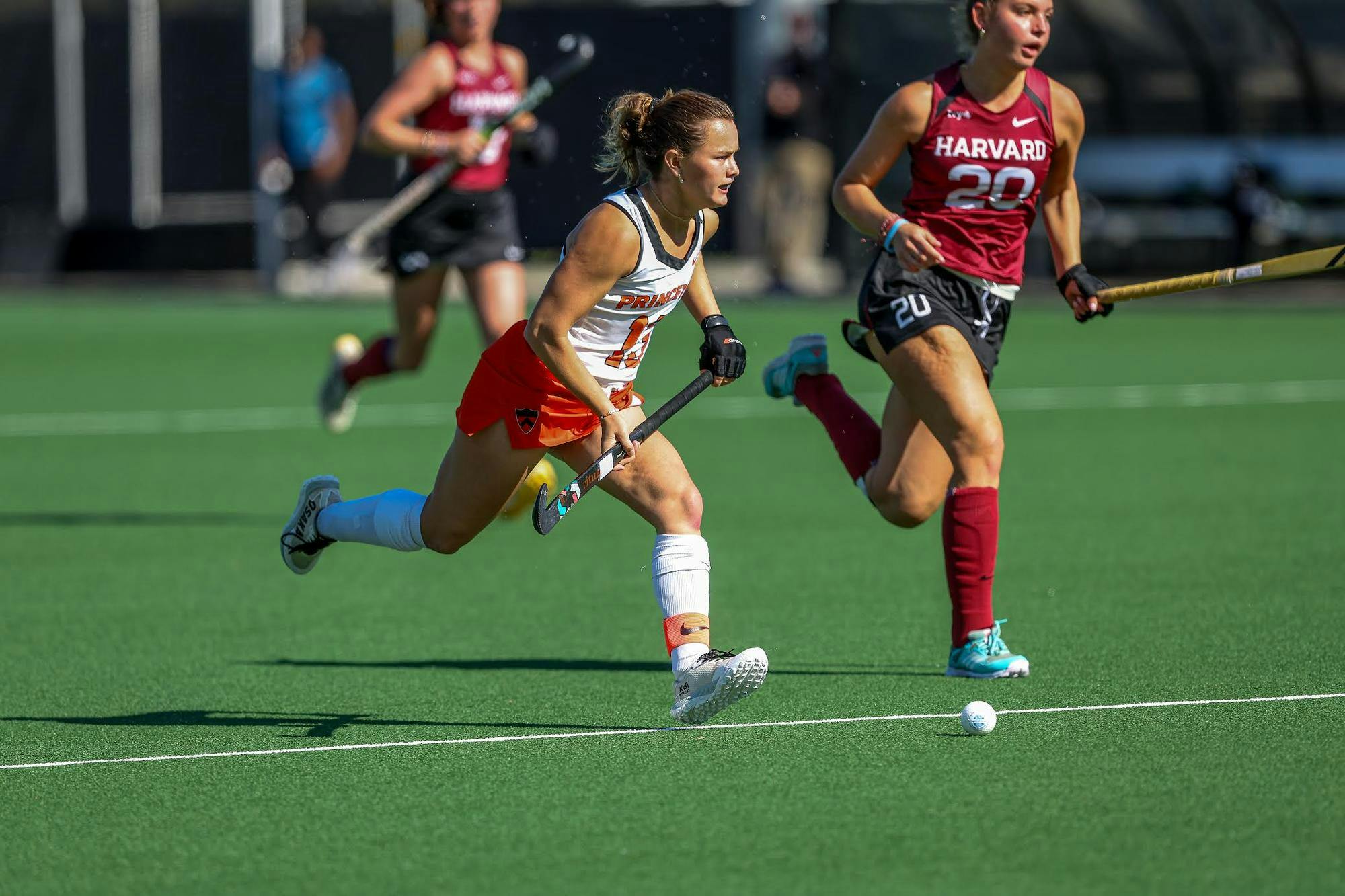A woman wearing an orange and white uniform on a grass field during a field hockey match with a stick in her hand. 