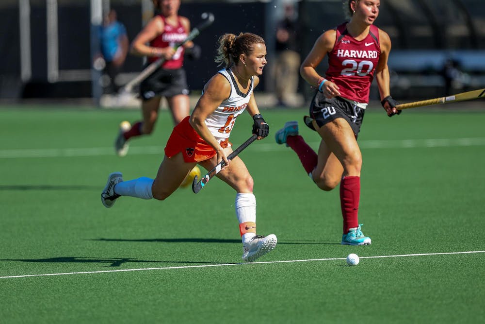 A woman wearing an orange and white uniform on a grass field during a field hockey match with a stick in her hand. 