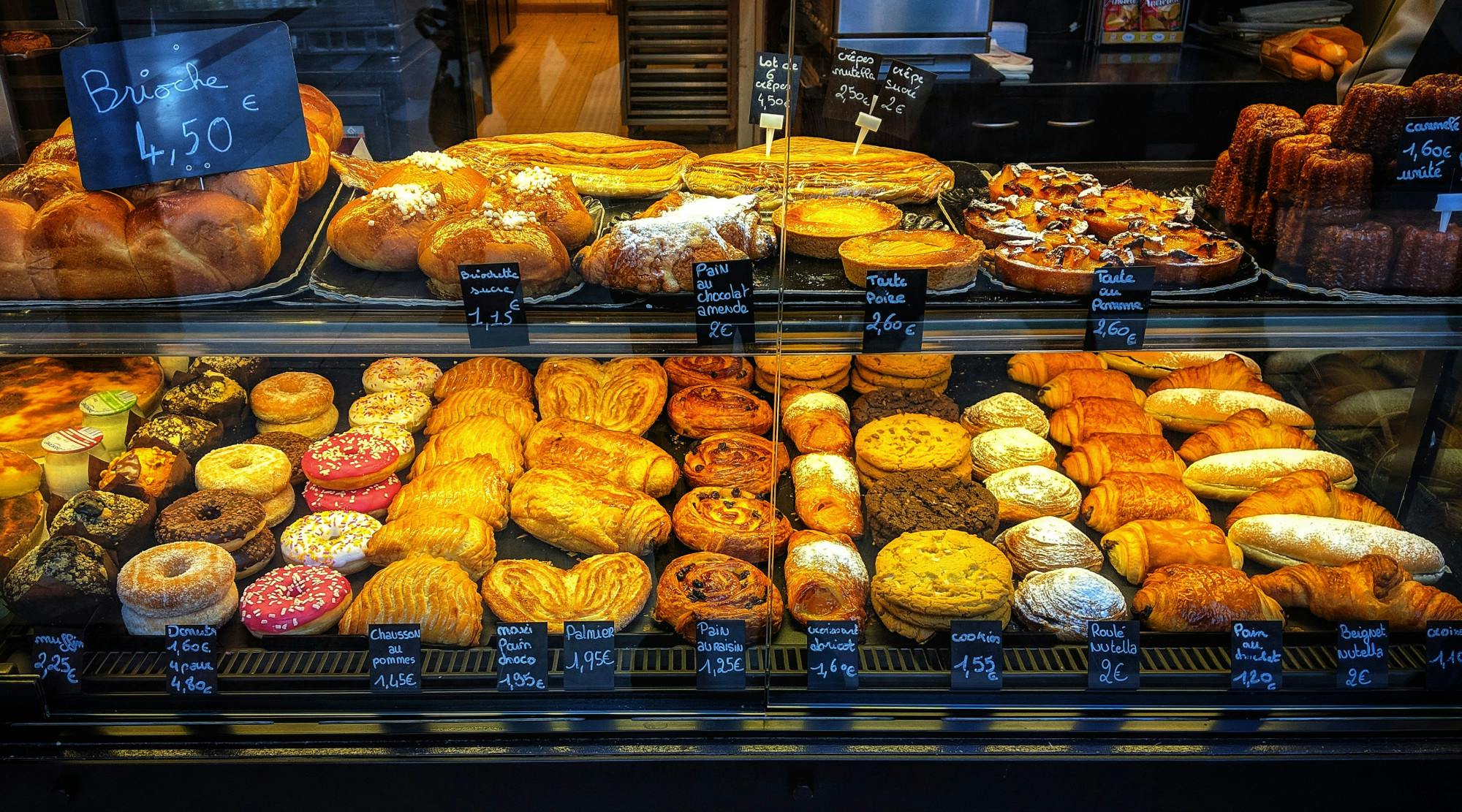 A colorful display of approximately thirty pastries behind a glass display case. The pastries are assembled in two rows and labeled with black labels. 
