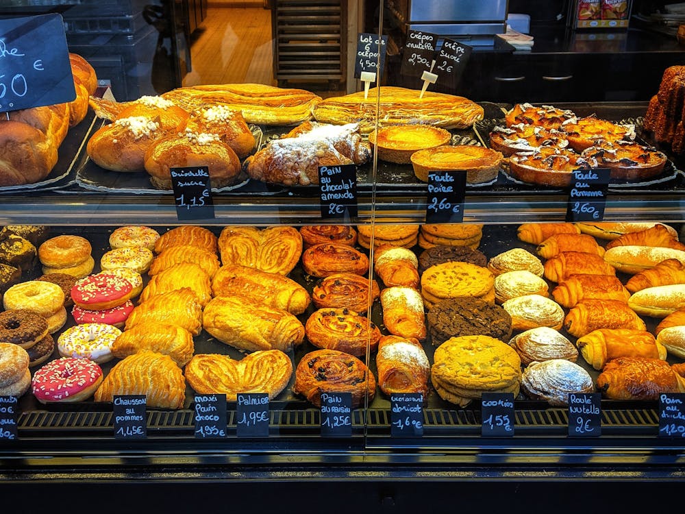 A colorful display of approximately thirty pastries behind a glass display case. The pastries are assembled in two rows and labeled with black labels. 