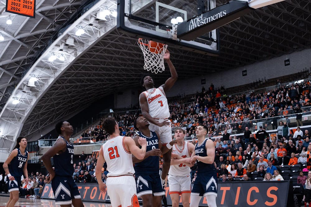 A basketball player in a white Princeton jersey dunks a basketball into the basket while players in Yale and Princeton jerseys watch from below