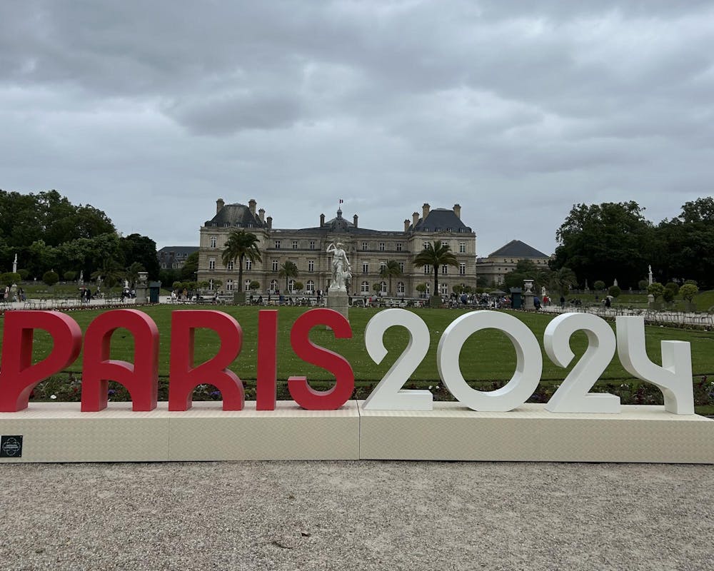 A large “Paris 2024” sign in bold red and white letters stands against a backdrop of a green lawn and a large historic building. There is a cloudy sky. 
