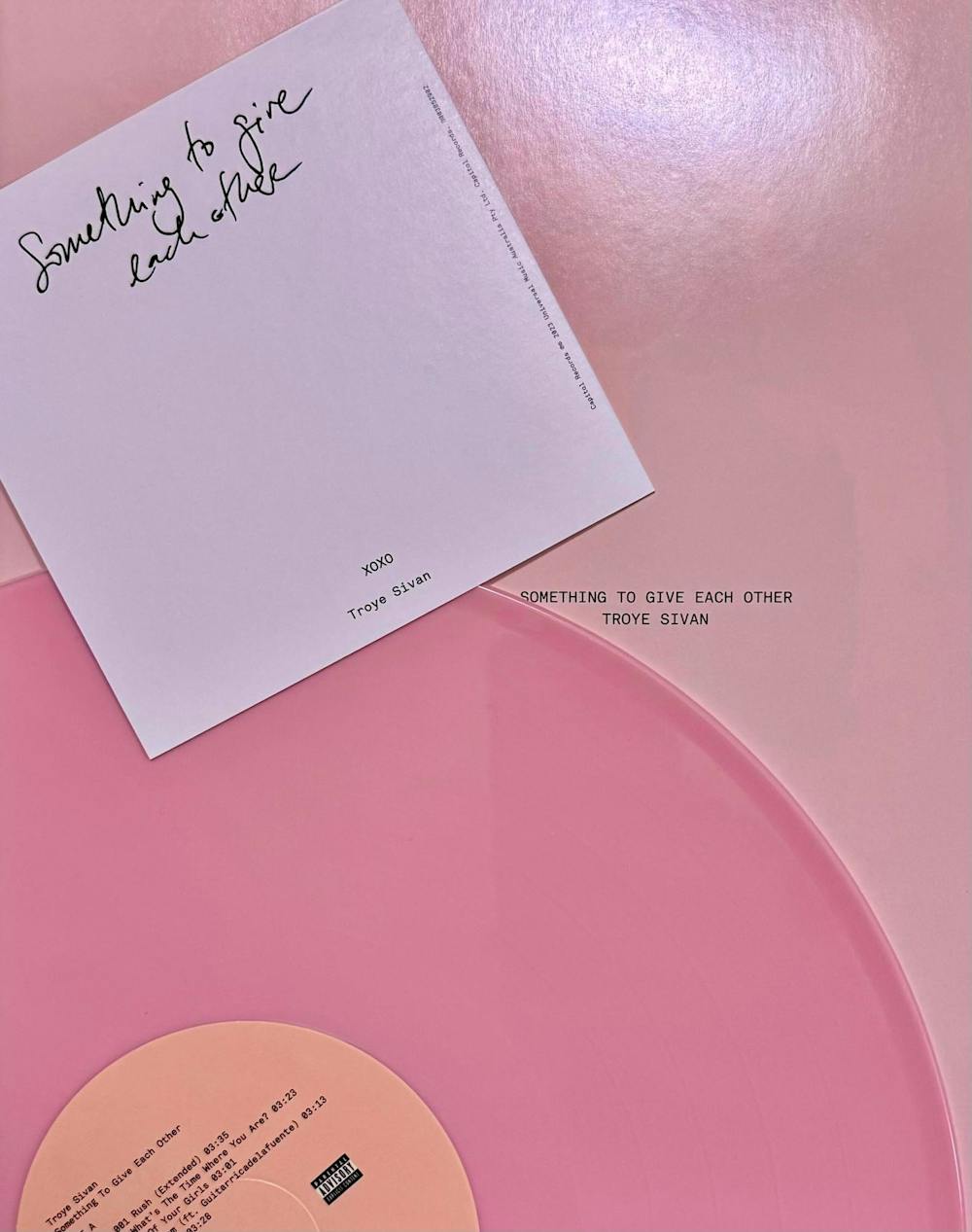 The signed vinyl cover of Troye Sivan's new album called "Something To Give Each Other." Part of hot pink vinyl is visible on a light pink background. On top of the vinyl is a lavender rectangle with the words "XOXO Troye Sivan" on it. The album title is on the light pink background. 