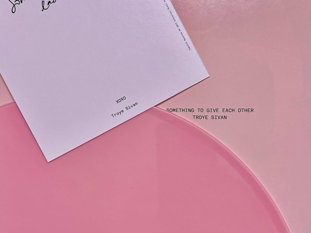 The signed vinyl cover of Troye Sivan's new album called "Something To Give Each Other." Part of hot pink vinyl is visible on a light pink background. On top of the vinyl is a lavender rectangle with the words "XOXO Troye Sivan" on it. The album title is on the light pink background. 