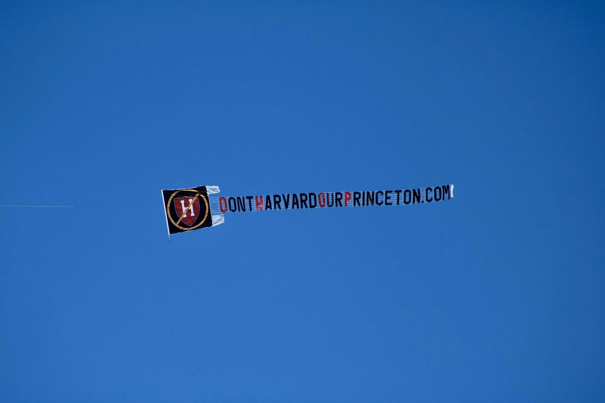 A banner flying through the sky on a sunny day. The banner reads DontHarvardourPrinceton.com