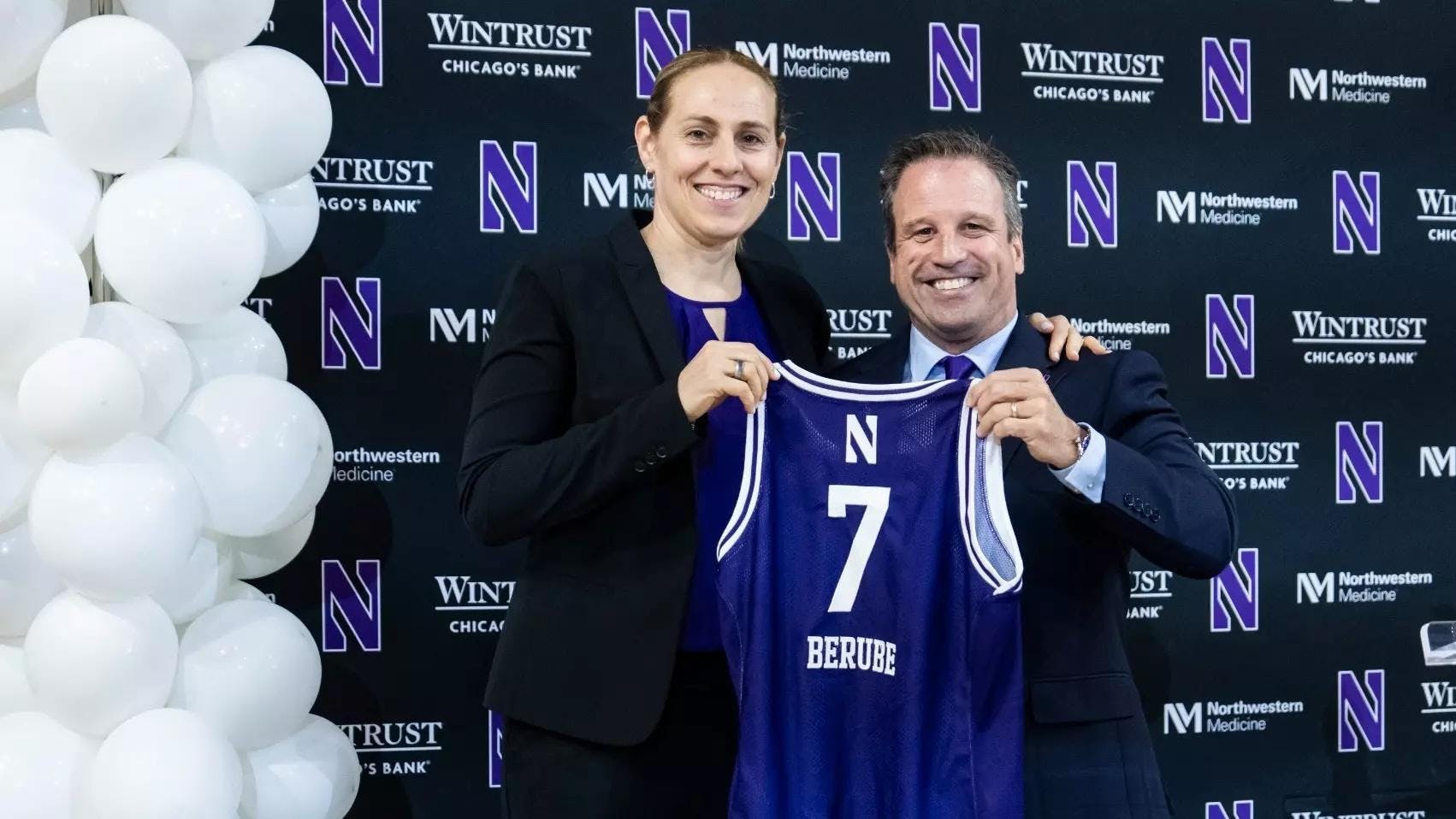 A woman in formal attire stands with a man in a suit, who holds a purple No. 7 jersey with the last name “Berube” during an introductory press event, with balloons in the background.