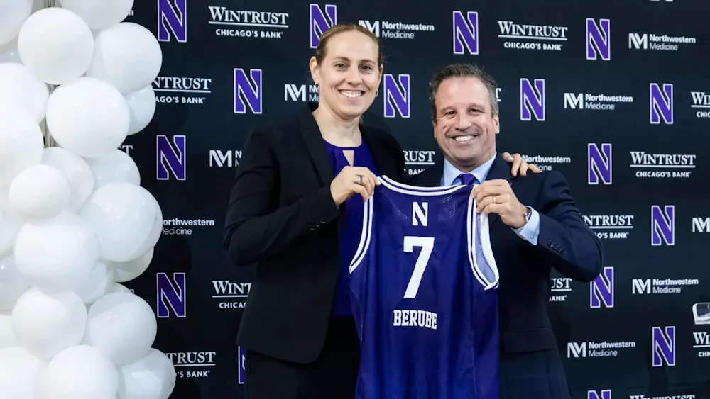 A woman in formal attire stands with a man in a suit, who holds a purple No. 7 jersey with the last name “Berube” during an introductory press event, with balloons in the background.