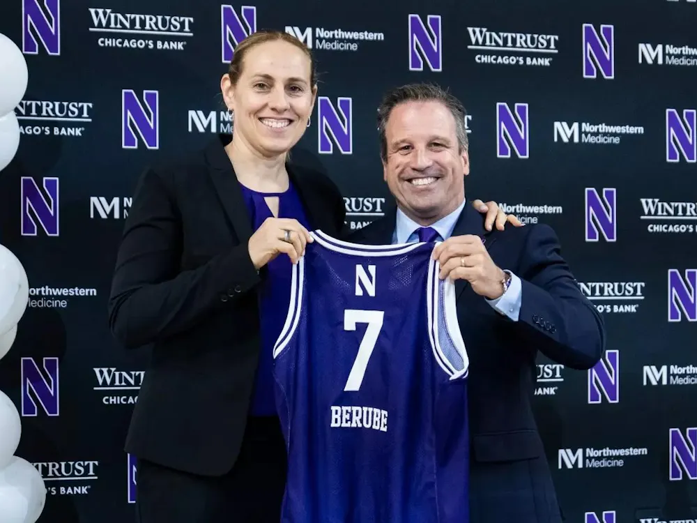 A woman in formal attire stands with a man in a suit, who holds a purple No. 7 jersey with the last name “Berube” during an introductory press event, with balloons in the background.