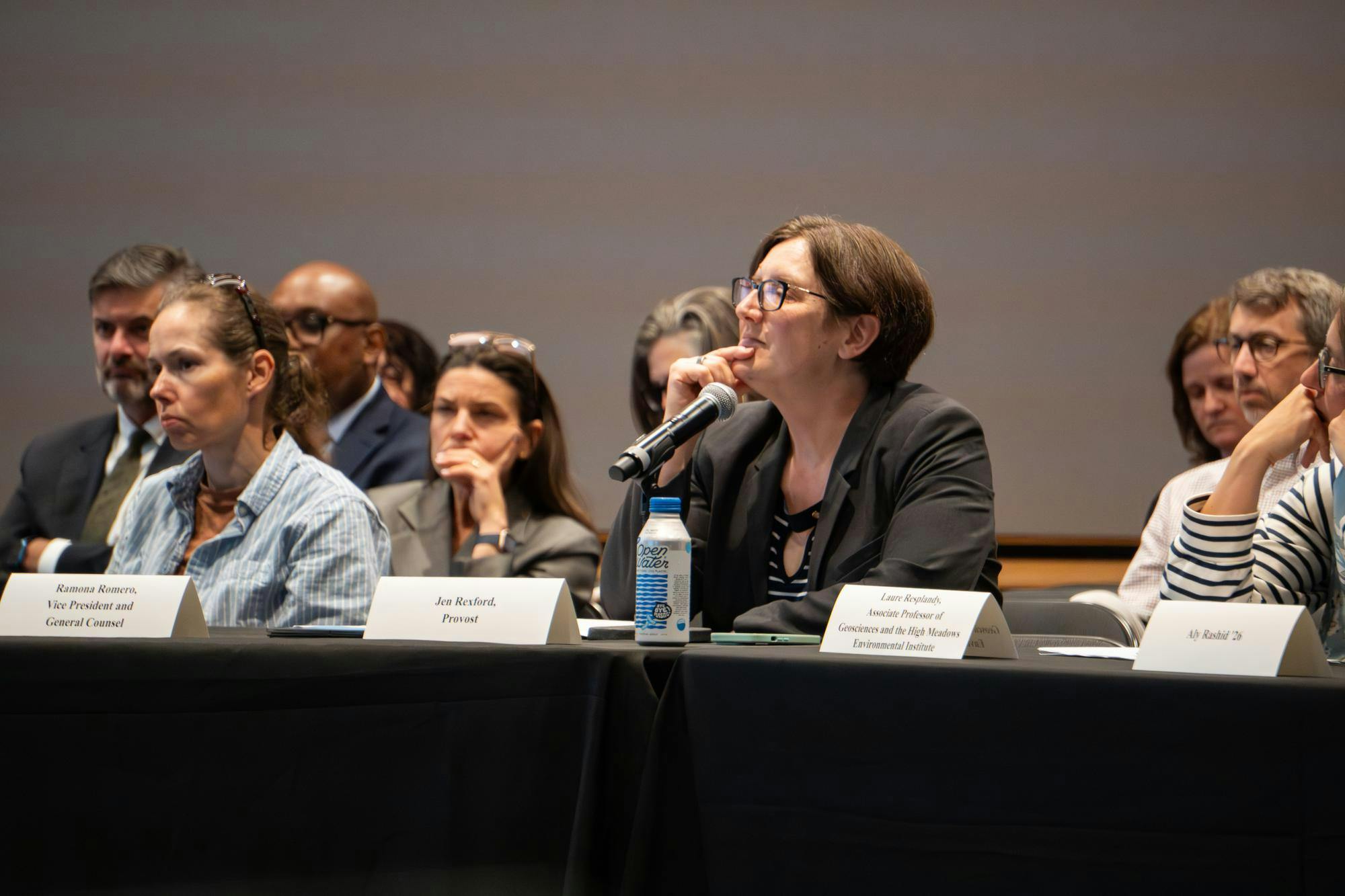 A group of people sit behind a black table with white name cards. A microphone sits in front of a woman with short brown hair and a dark grey blazer.