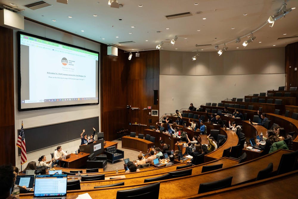 Large lecture hall with wood paneling and arced tables populated by students, some of which are raising their hands.