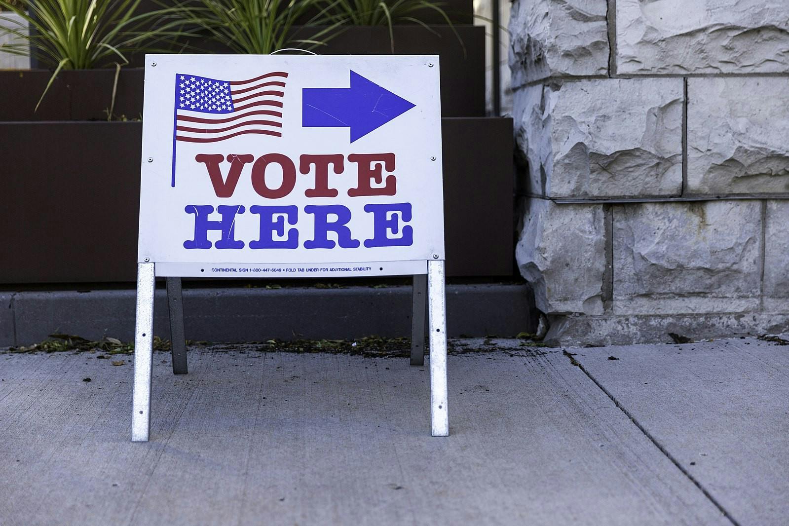 A sign in front of a planter and rock wall. The sign says “VOTE HERE,” with an American flag and arrow pointing to the left above.