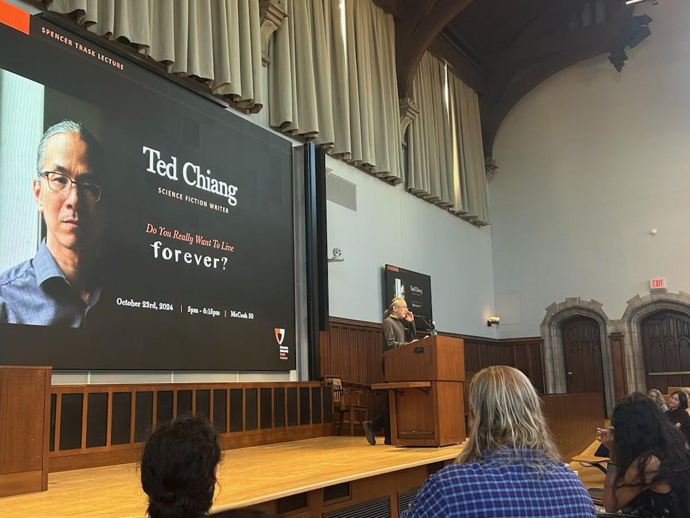 A person with glasses and a ponytail stands at a wooden podium on a stage, in front of a projection reading, "Ted Chiang, science fiction writer, Do you really want to live forever?