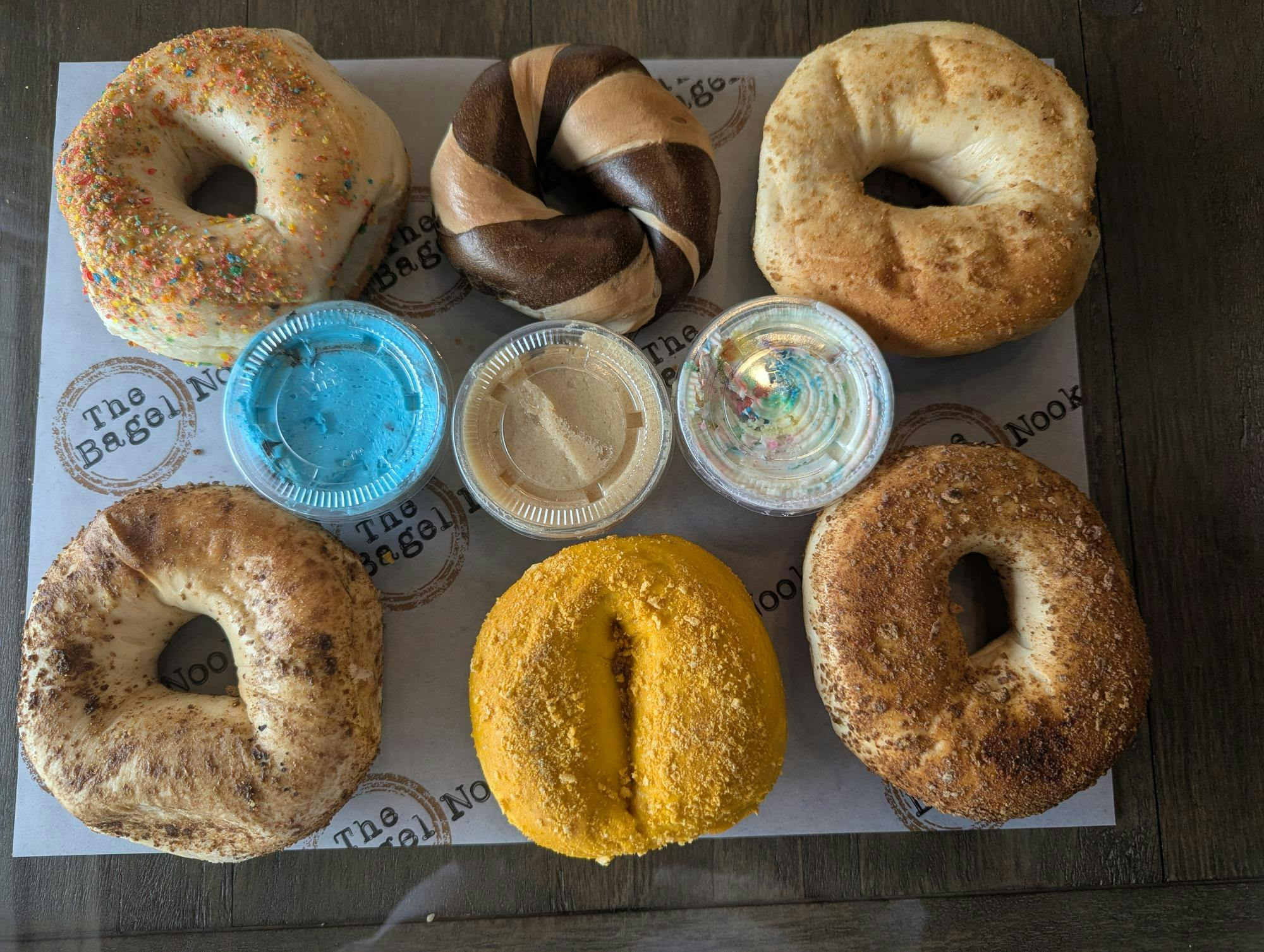 An assortment of bagels on parchment paper labeled "The Bagel Nook." The bagels are divided into two rows, each row containing three bagels. Between the rows are three plastic containers of bagel spreads.