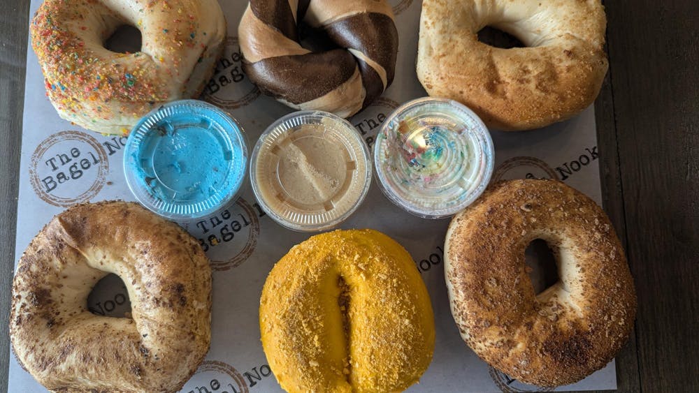 An assortment of bagels on parchment paper labeled "The Bagel Nook." The bagels are divided into two rows, each row containing three bagels. Between the rows are three plastic containers of bagel spreads.