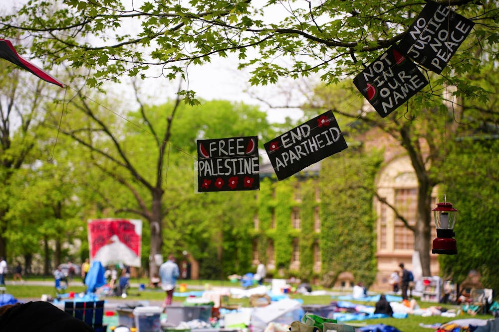 Signs reading "Free Palestine," "End apartheid," "Not in our name," and "No justice no peace" hang from a tree in the foreground. In the background, people sitting on a green lawn and a stone building covered in ivy. 