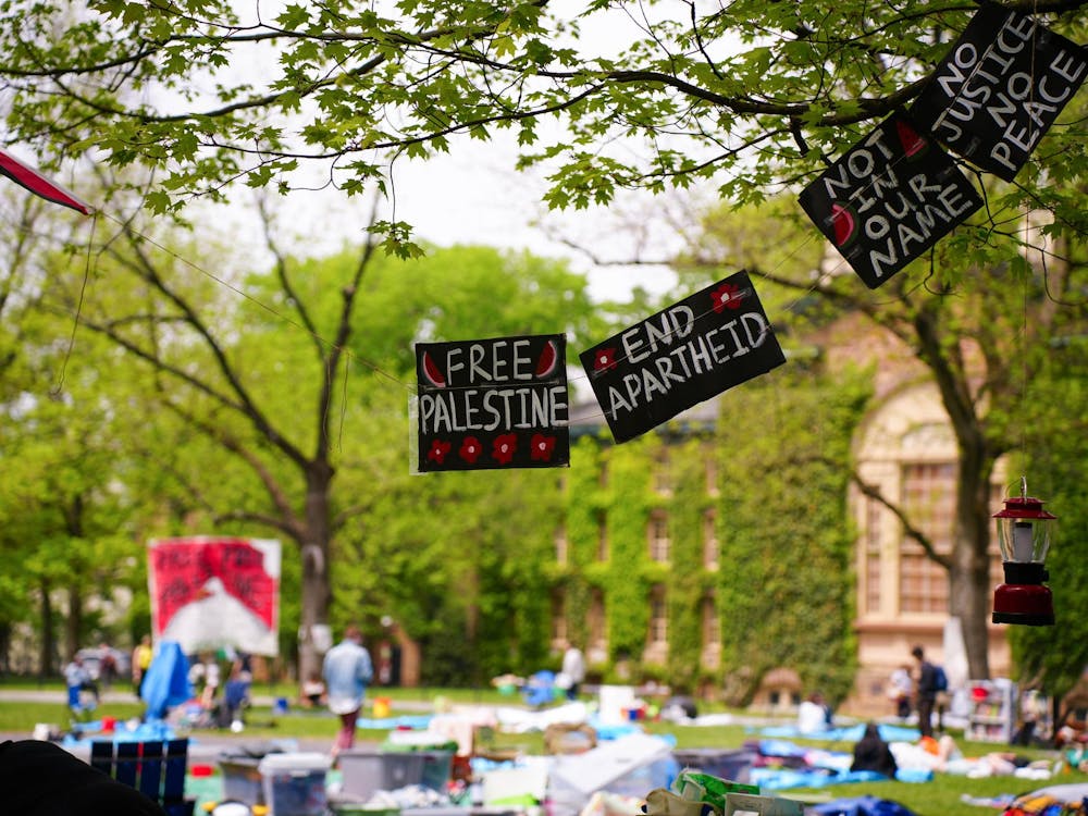 Signs reading "Free Palestine," "End apartheid," "Not in our name," and "No justice no peace" hang from a tree in the foreground. In the background, people sitting on a green lawn and a stone building covered in ivy.