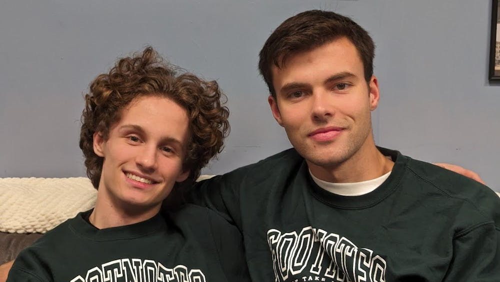 Two people sit on a couch wearing green sweatshirts that say “Footnotes.” 