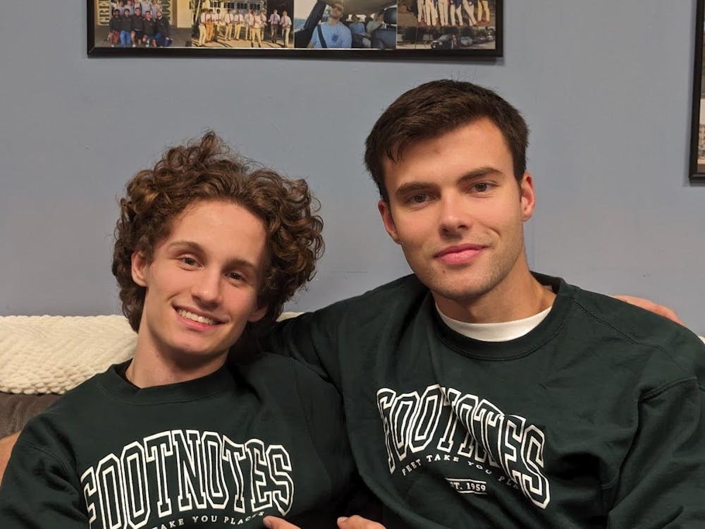 Two people sit on a couch wearing green sweatshirts that say “Footnotes.” 