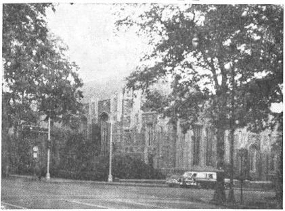 A black and white photo of Madison Hall from the corner of Nassau Street and University Place. An image of a gothic-style building behind trees and lamp posts.