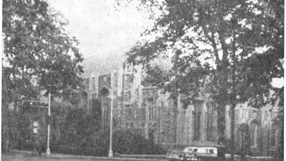 A black and white photo of Madison Hall from the corner of Nassau Street and University Place. An image of a gothic-style building behind trees and lamp posts.