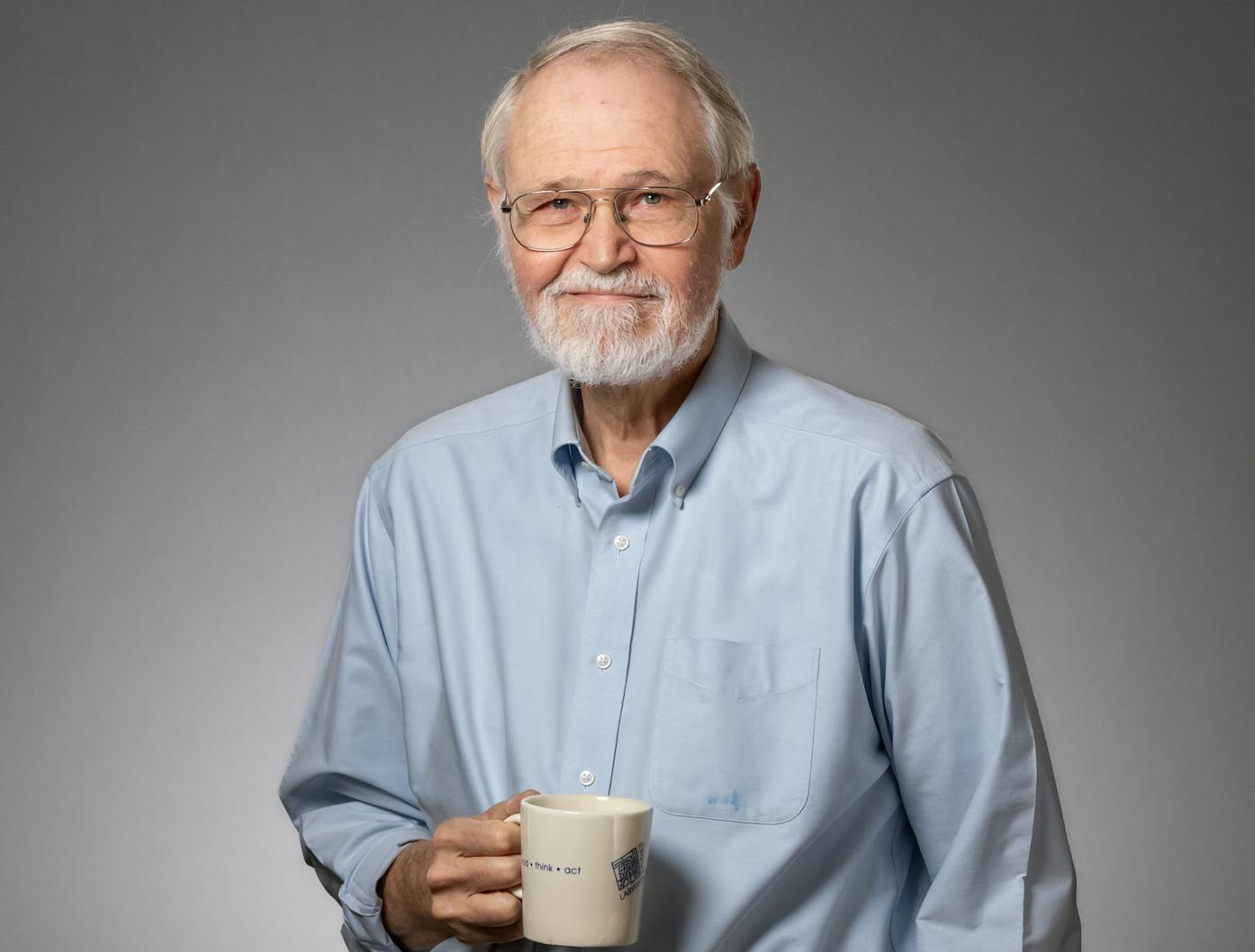 A man with silver hair and beard, blue eyes, and glasses smiles at the camera. He wears a light blue button-up and holds a white mug in his hand. 