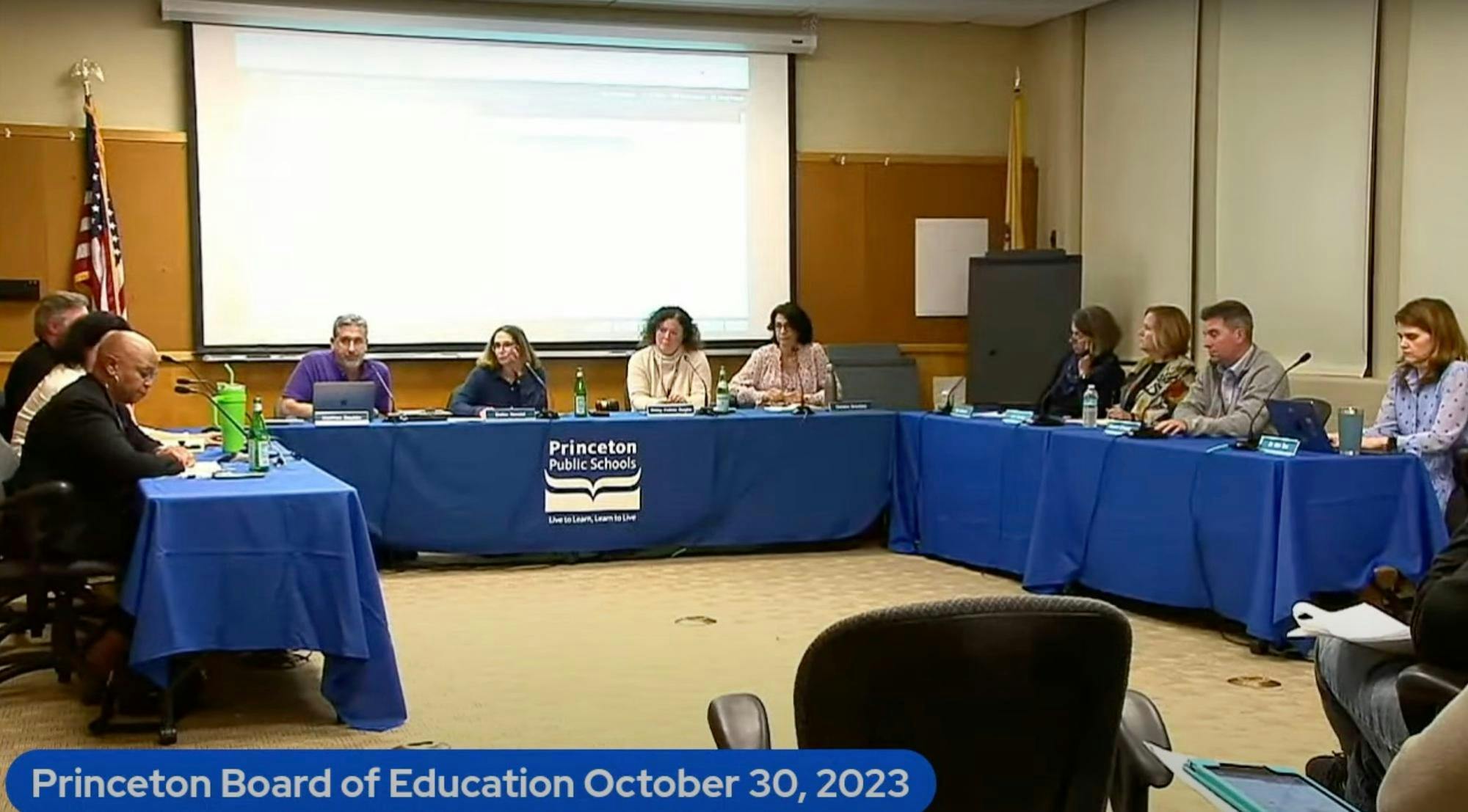 A group of people sit behind a table in a meeting room, under an illuminated projector screen.