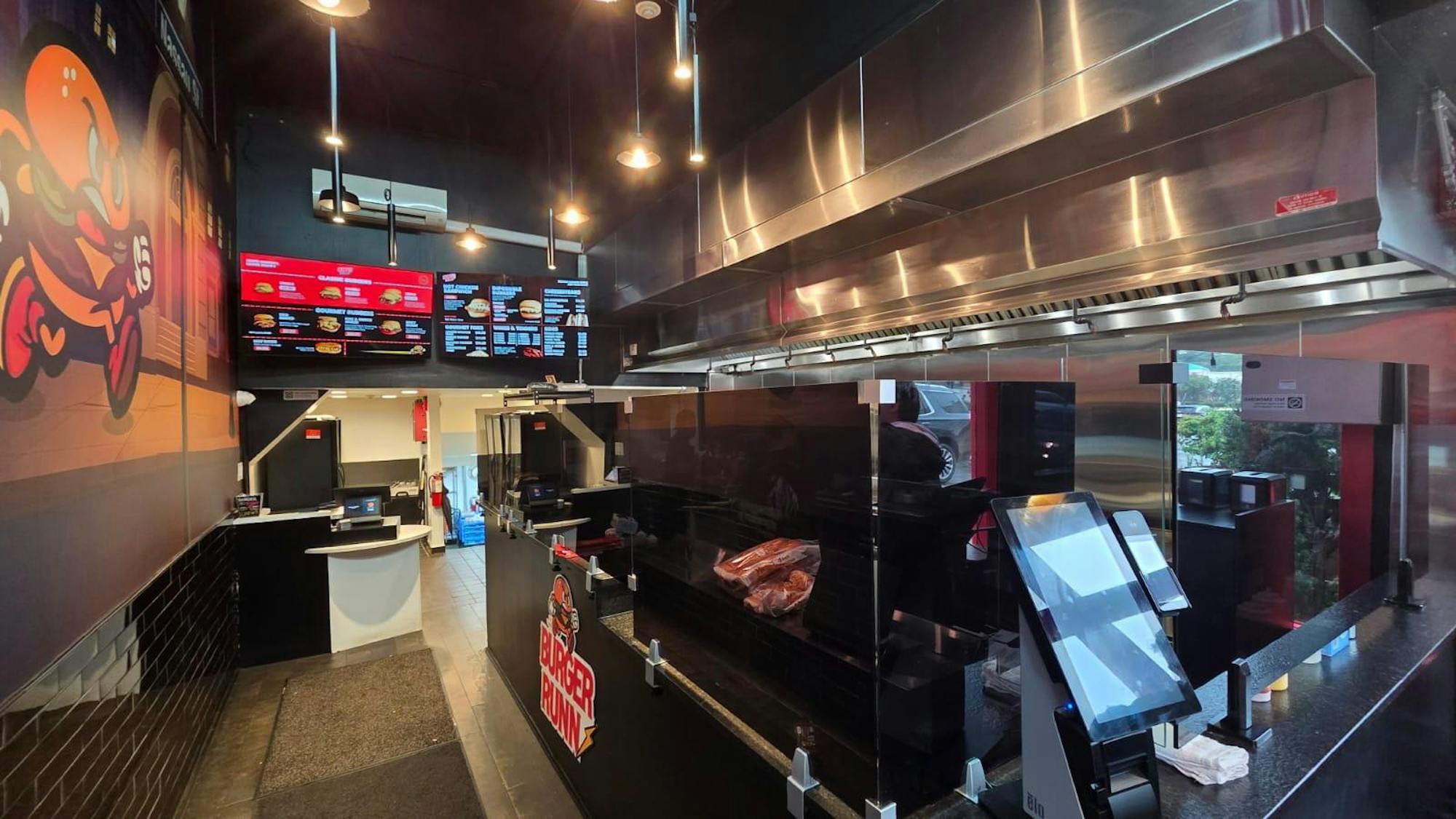 The inside of the restaurant has black walls and a black counter. Part of a kitchen is visible with silver heat vents. There are two TVs displaying the BurgerRunn menu on it.