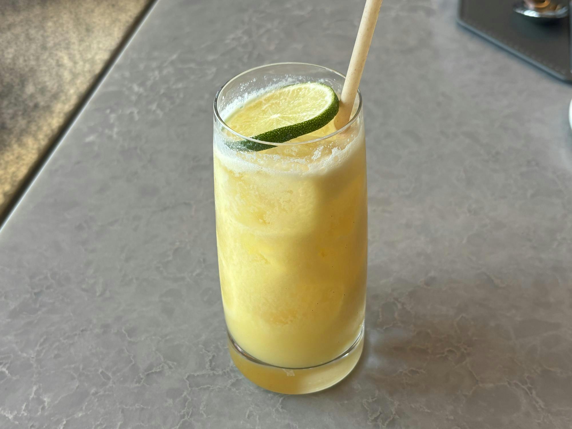 A yellow drink is in a clear glass with a slice of lime and a light brown straw.