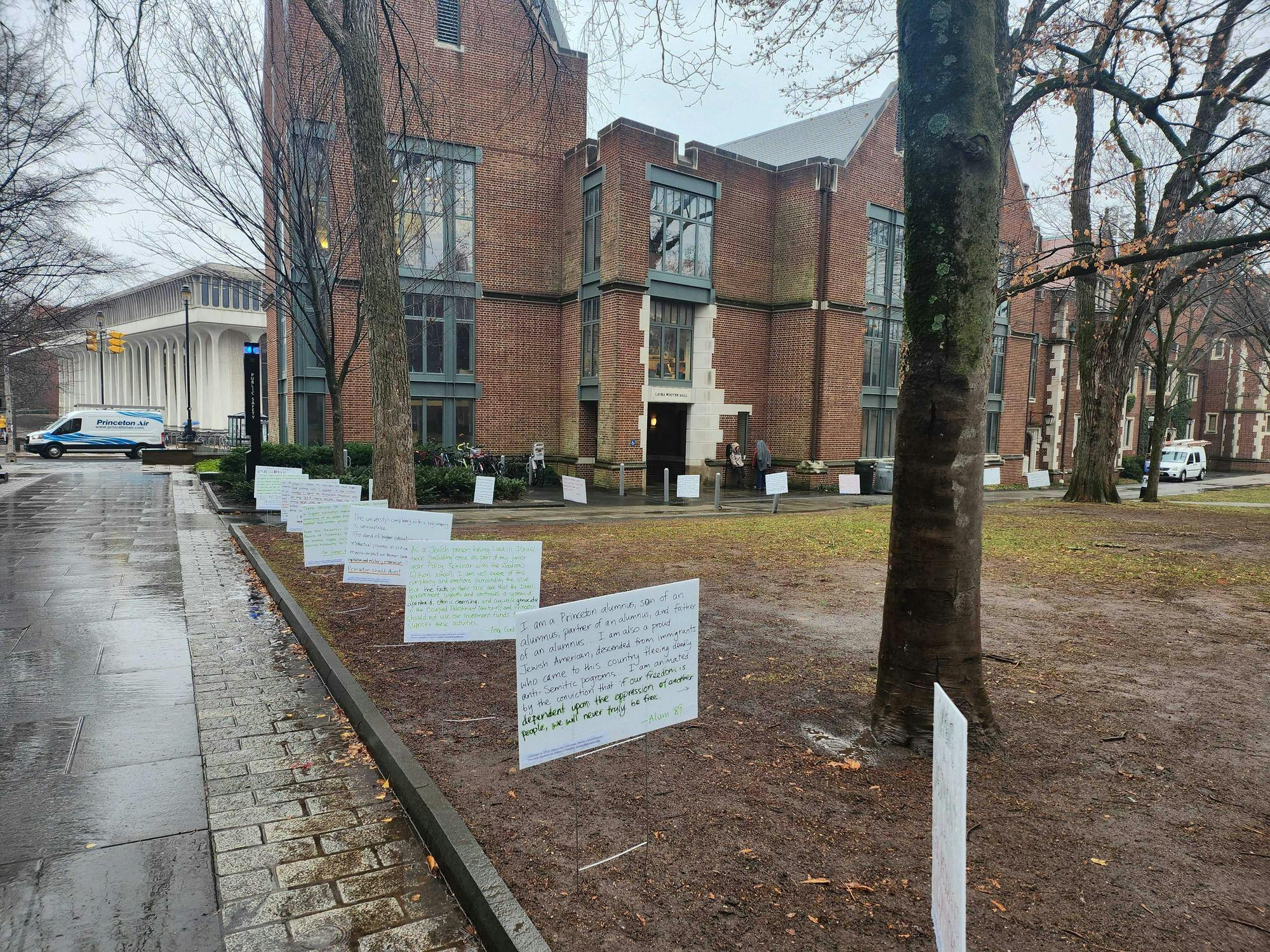 Poster board with writing are planted into a lawn. The lawn sits in front of a brick building and next to a sidewalk. It is raining. 