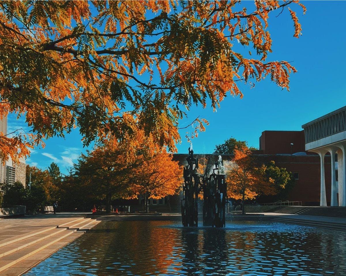 blue sky in front of a wide area with stone floors and steps and a fountain with water coming out of it.  There are also buildings at the back.