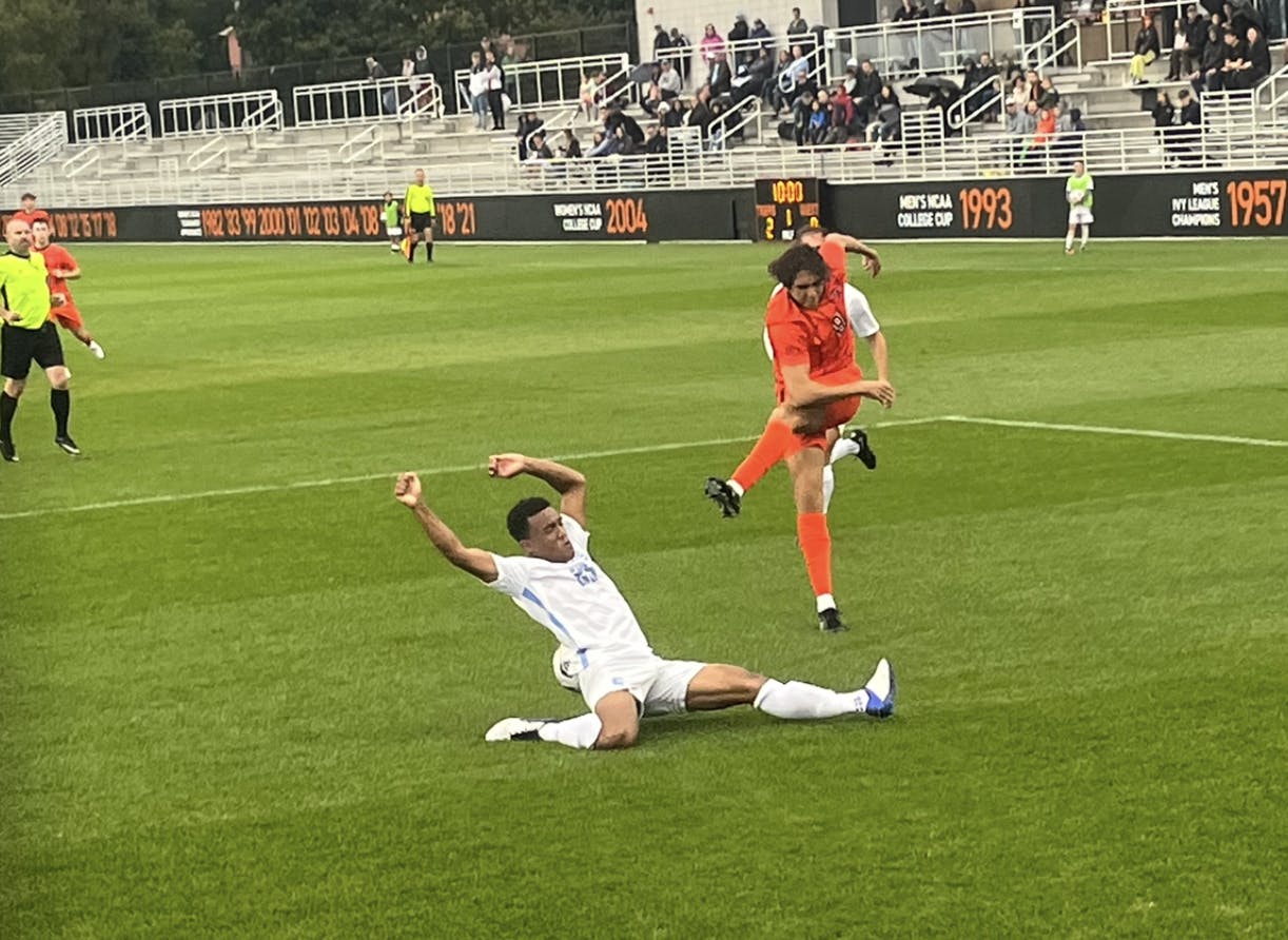 Man in orange kicks ball with right leg over a sliding man in white with legs stretched. 