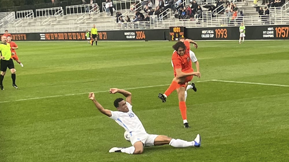 Man in orange kicks ball with right leg over a sliding man in white with legs stretched. 
