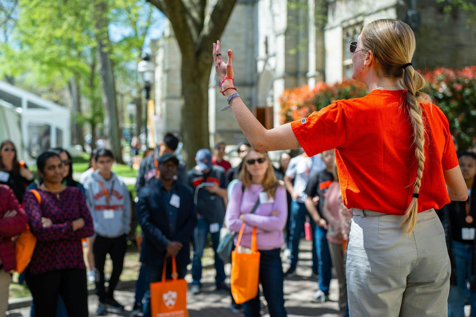 A student with a blonde braid dressed in an orange t-shirt addresses a crowd of adults and high-school aged students, with trees and a white tent in the background