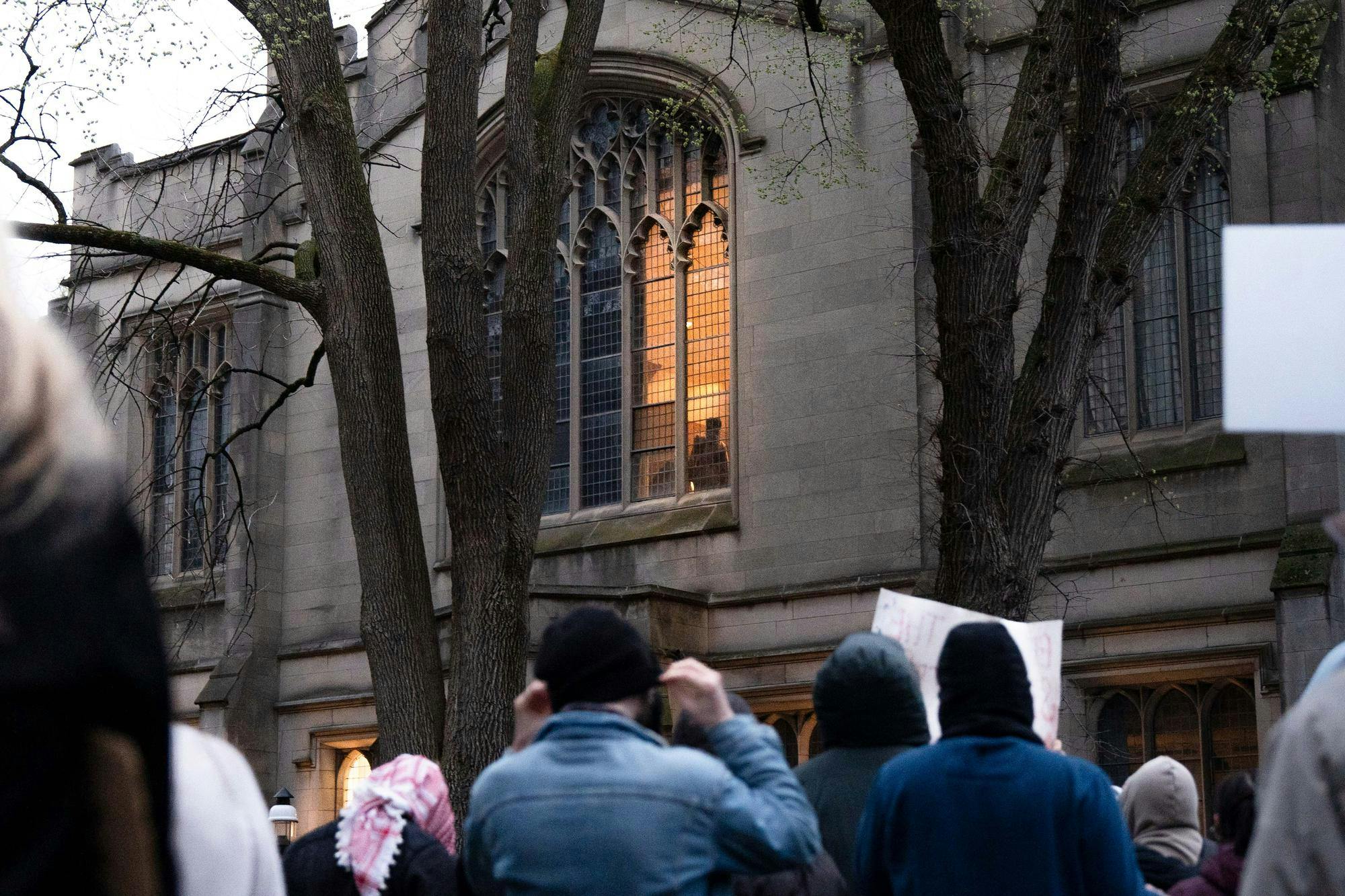 In the foreground, a group of protesters holds signs. Beyond the protesters is a large gothic building with a lit window, framed by trees. Inside the window, an orange light  illuminates the silhouette of two figures. 