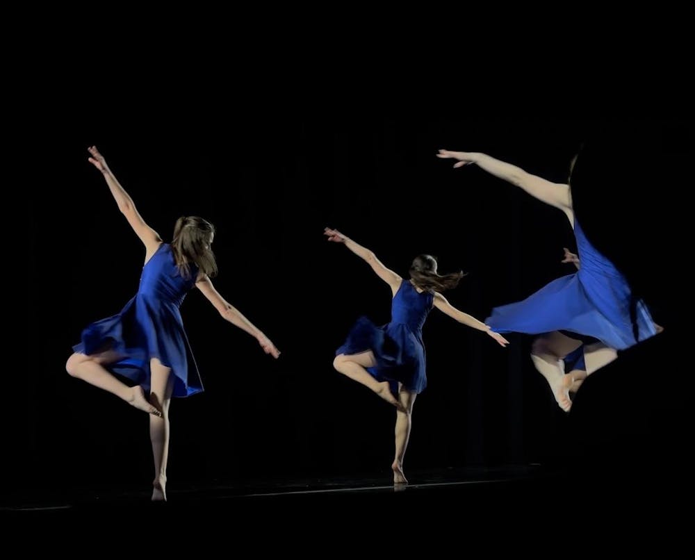 Three dancers in blue dresses do a turn on one leg.