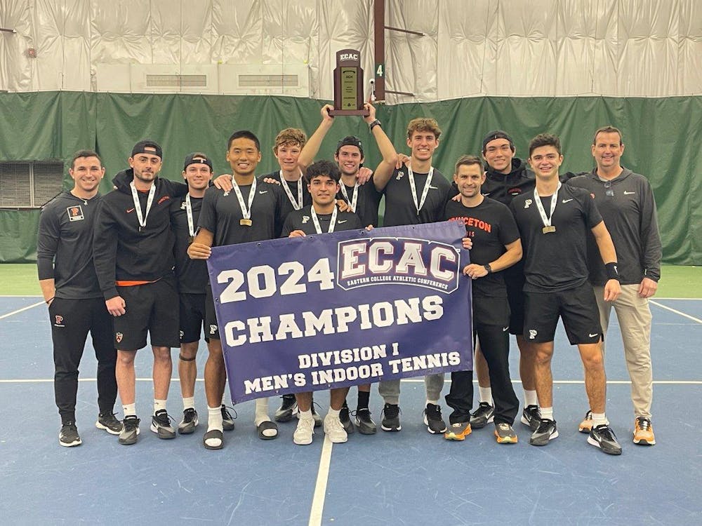 Men in black shirts smile for a picture, standing on a tennis court holding a banner and a trophy.