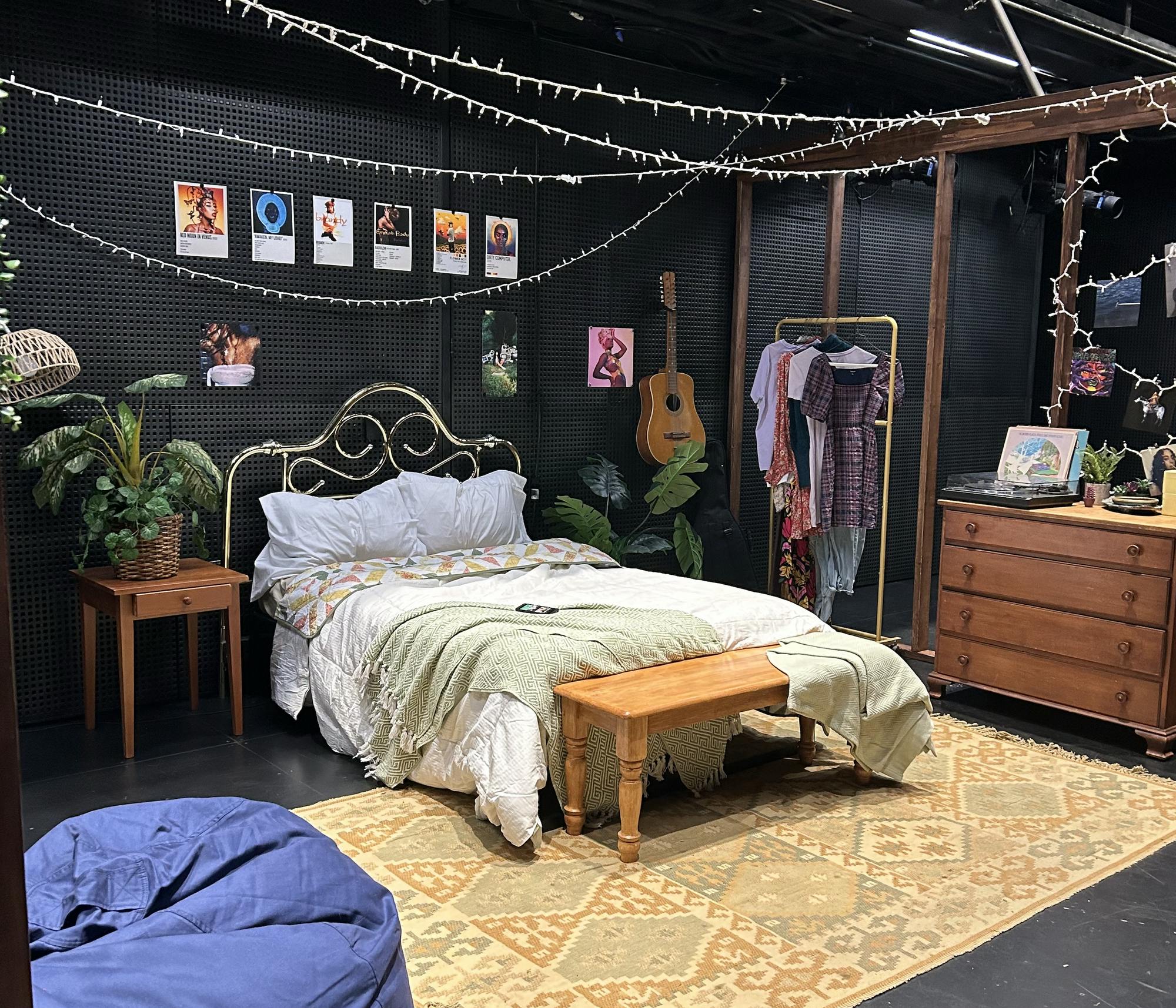 A bed with fluffy green bedsheets and two pillows at the center of a black room, surrounded by decorative features like album covers, carpets, drawers, and plants.