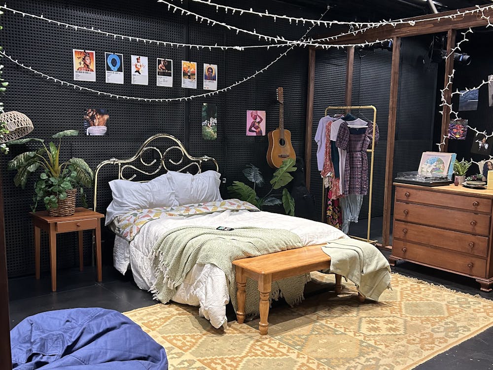 A bed with fluffy green bedsheets and two pillows at the center of a black room, surrounded by decorative features like album covers, carpets, drawers, and plants.