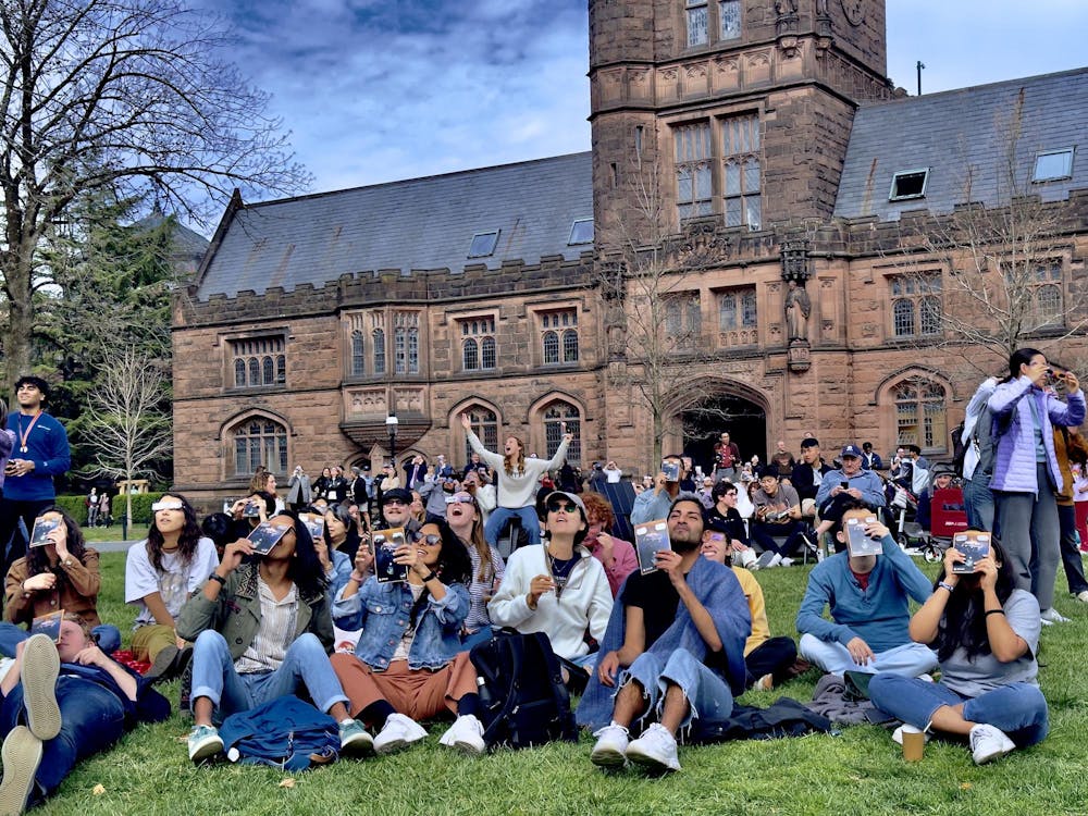 A large crowd of students sit on a patch of green grass and look up at the sky with an old, brown stone building behind them.
