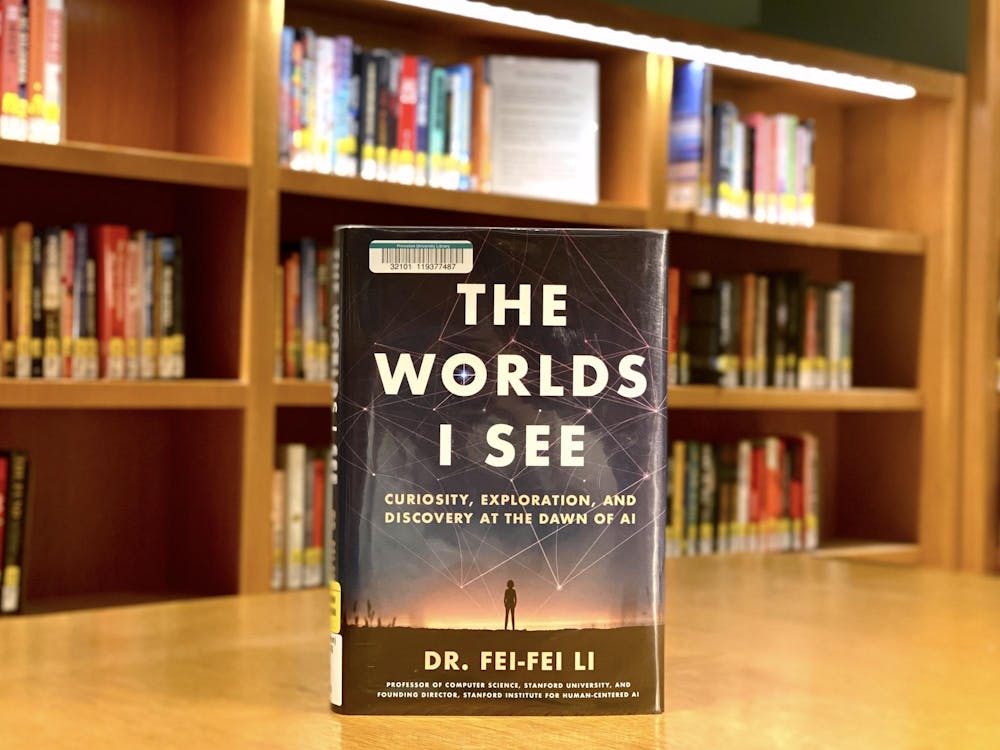 "The Worlds I See" by Dr. Fei Fei Li standing up on a tan library table, with bookshelves in the background.