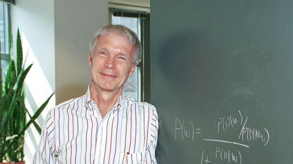 An elderly man with white hair stands in front of a potted plant and a green chalkboard.