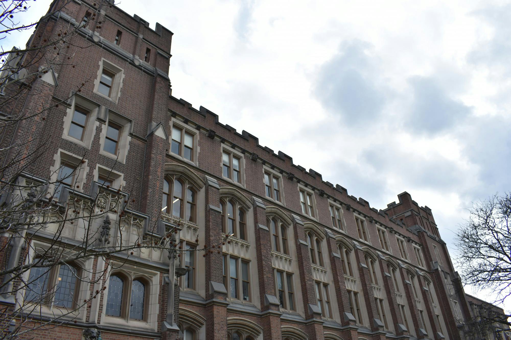 A Gothic style building featuring tall, narrow windows and made up of red brick with stone trim around the windows and corners. The sky behind is partly cloudy.