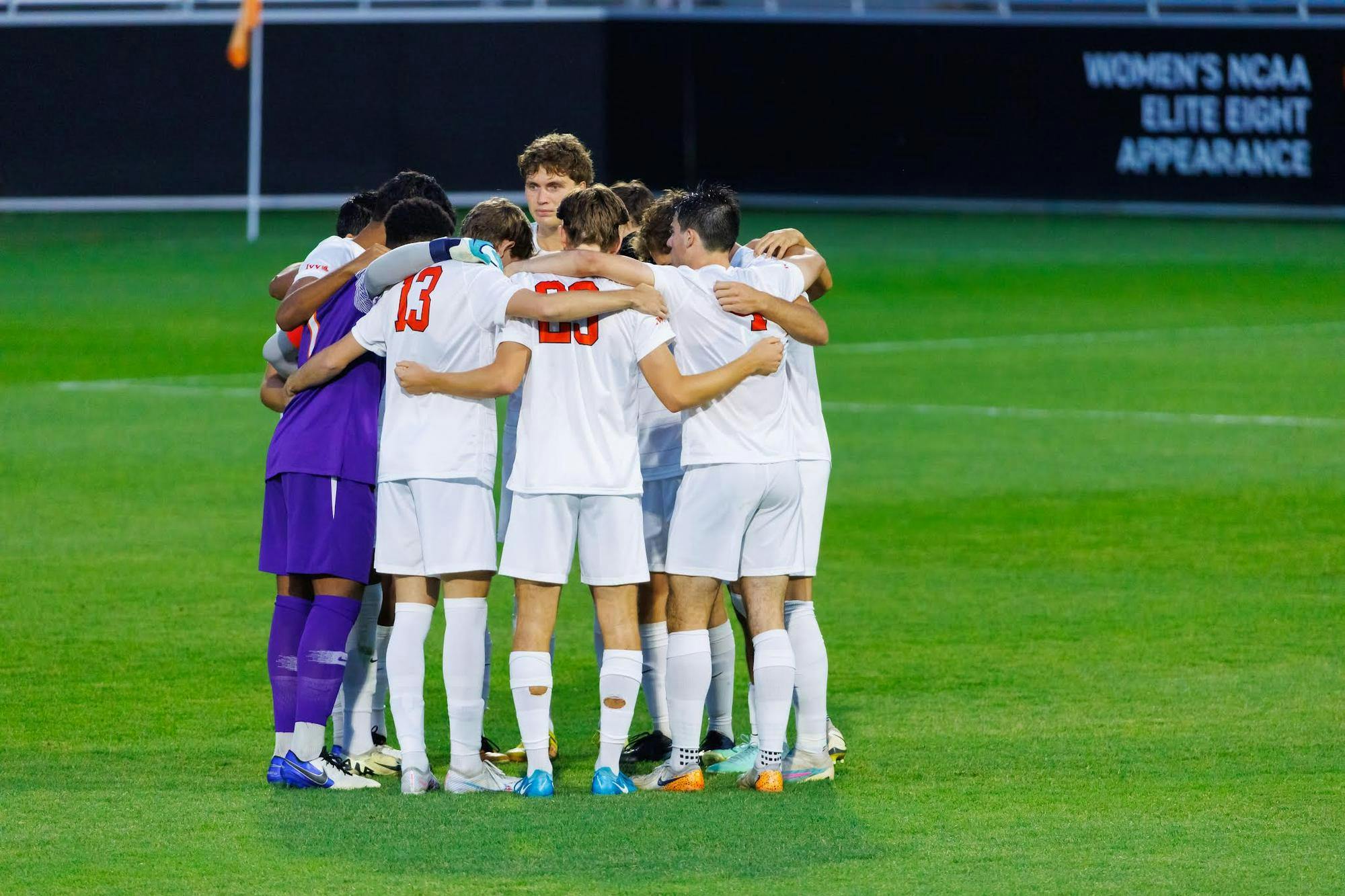 A group of men in a circle on a soccer field with arms around their shoulders. 