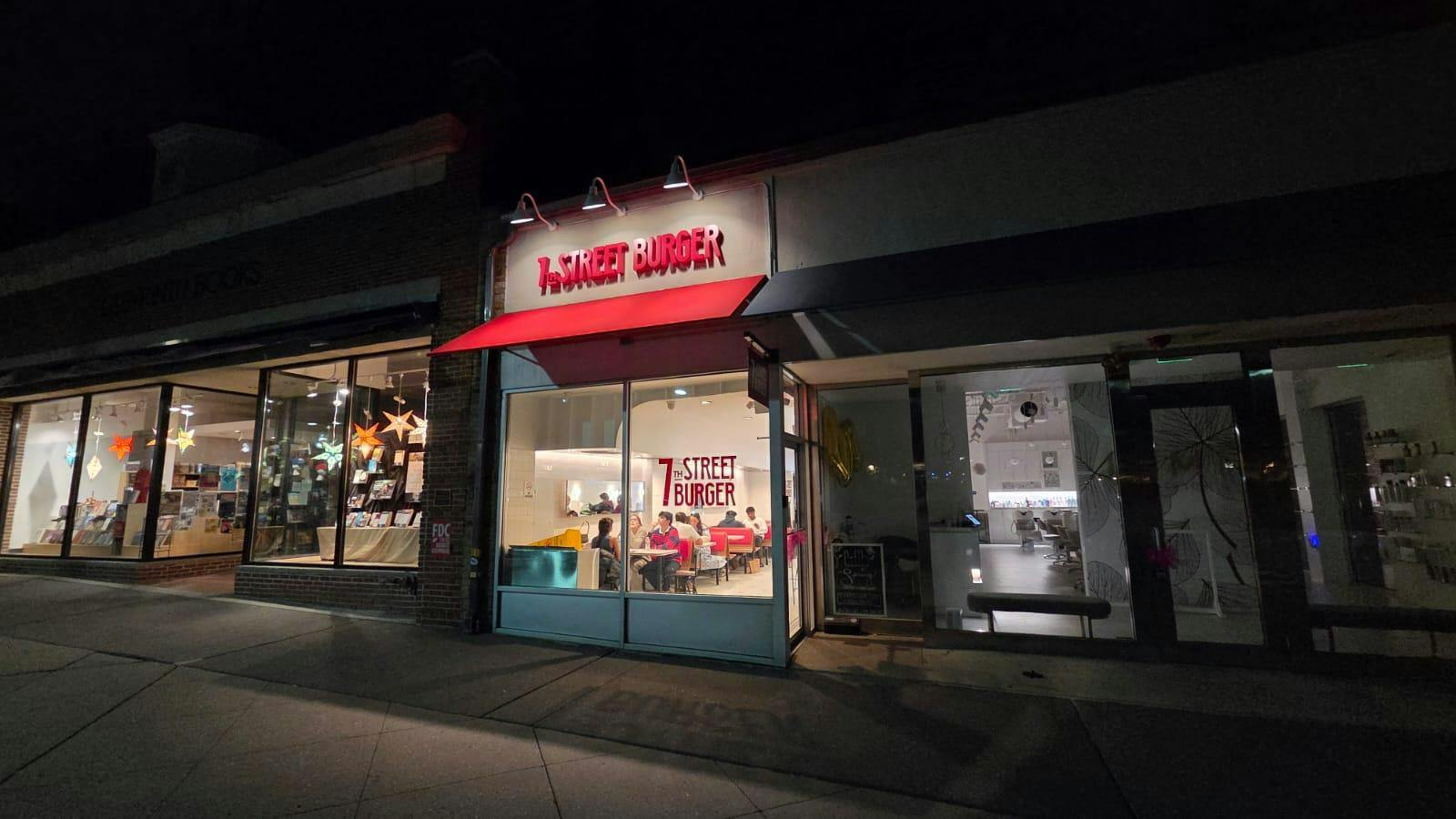 A row of stores with white exteriors lines a concrete sidewalk. One store is particularly lit up. It has a red awning with a red sign above it that reads "7th Street Burger." People sitting and eating a visible through the window.