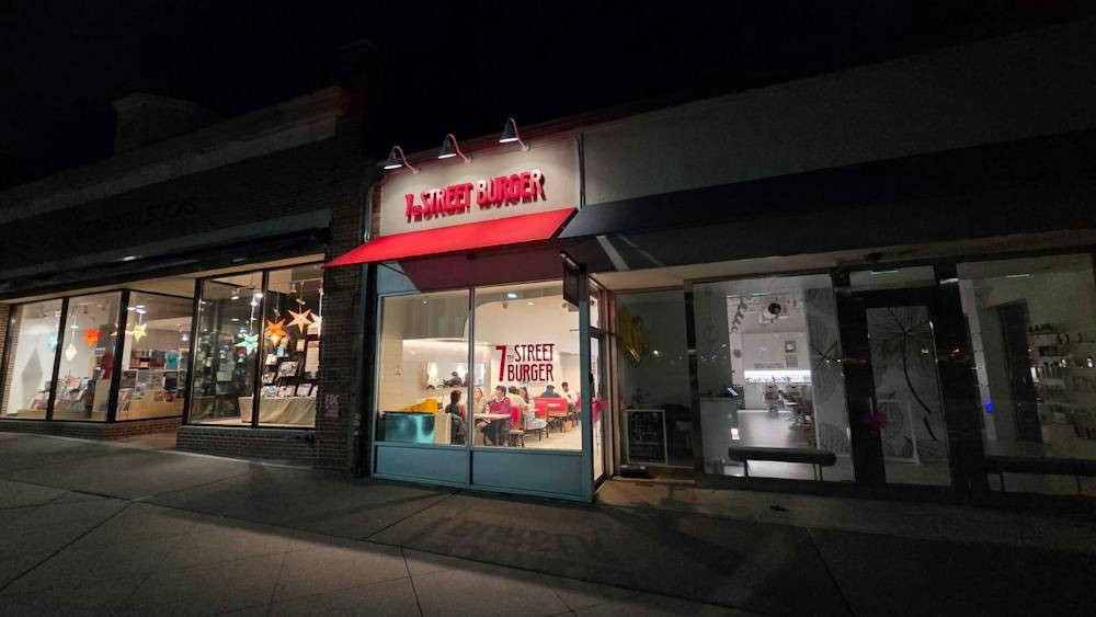A row of stores with white exteriors lines a concrete sidewalk. One store is particularly lit up. It has a red awning with a red sign above it that reads "7th Street Burger." People sitting and eating a visible through the window.