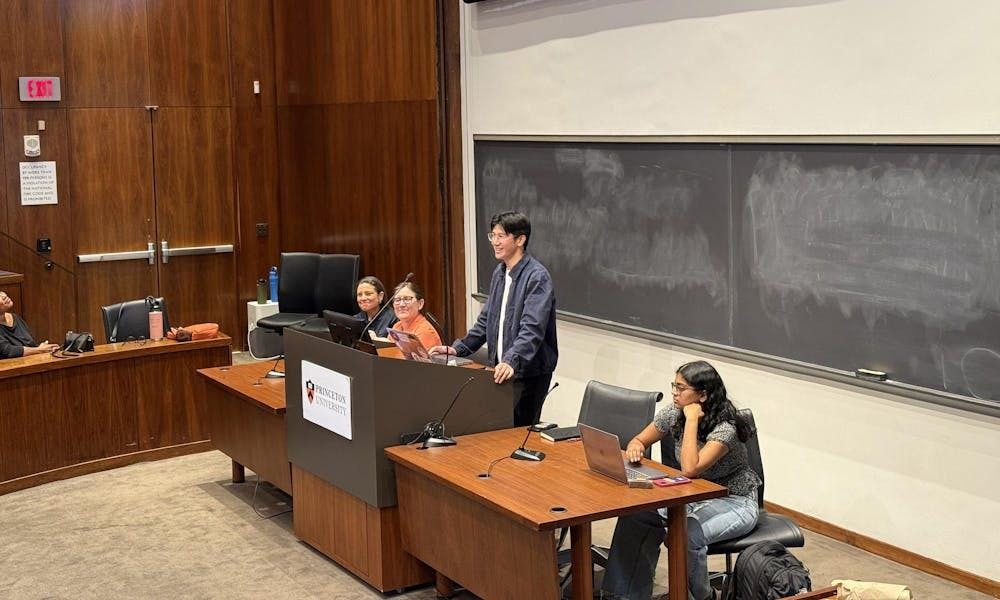 Cuatro personas están sentadas y de pie detrás de un escritorio en un auditorio. Hay una pizarra detrás de ellas.
