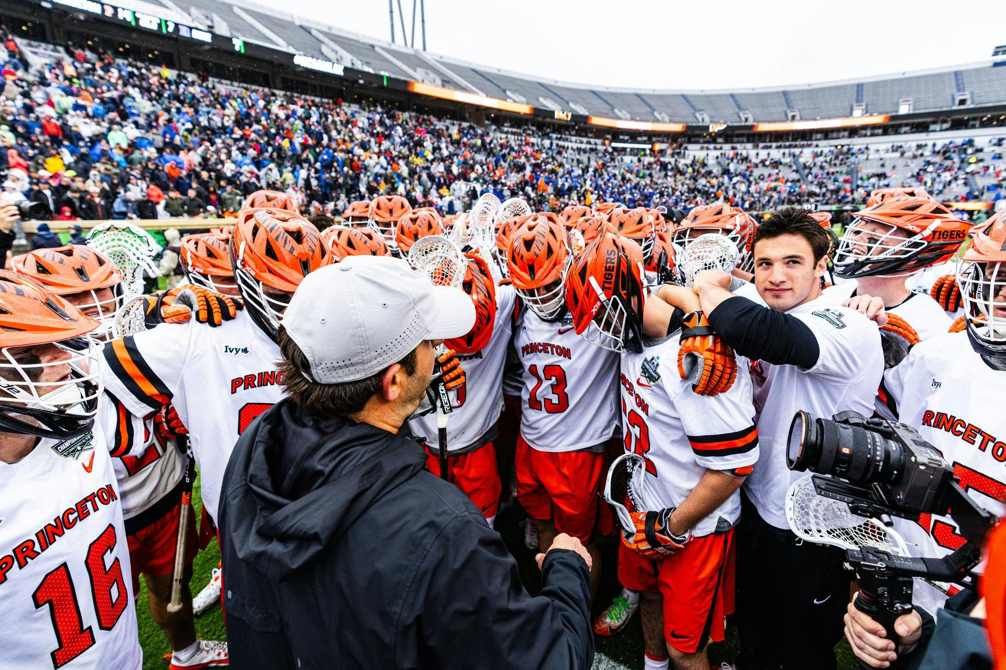 Princeton men’s lacrosse players huddle together on the field after a game, surrounded by fans in a stadium.