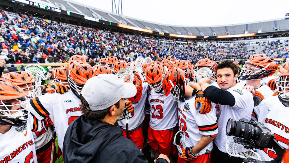 Princeton men’s lacrosse players huddle together on the field after a game, surrounded by fans in a stadium.