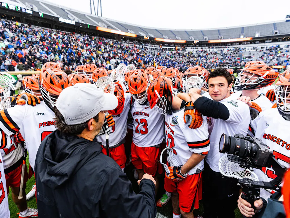 Princeton men’s lacrosse players huddle together on the field after a game, surrounded by fans in a stadium.