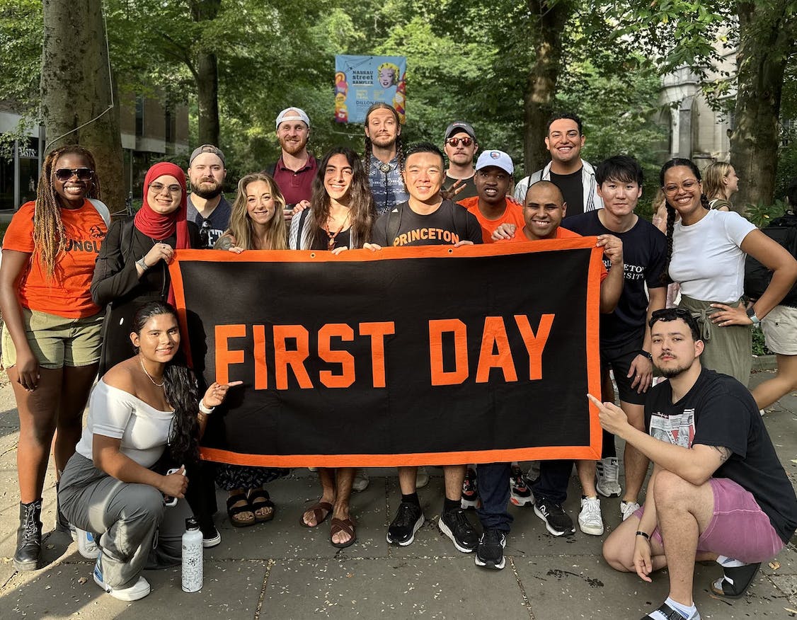 A group of transfer students stands by McCosh hall holding a flag that says "First Day" and smiling.